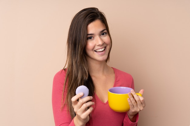 Teenager woman over isolated wall holding colorful French macarons and a cup of milk