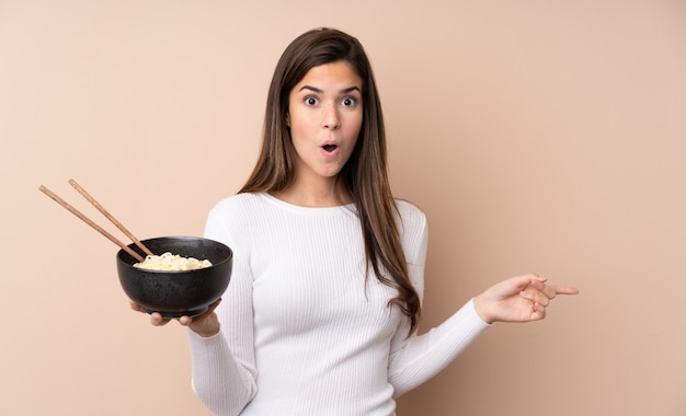 Teenager woman over isolated wall surprised and pointing side while holding a bowl of noodles with chopsticks