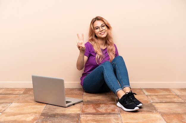 Teenager woman with a laptop sitting on the floor at indoors smiling and showing victory sign