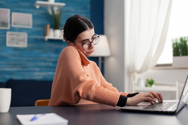 Teenager woman working at e-learning platform for university\
project during remote education. student talking at phone about\
digital courses while sitting at desk in living room