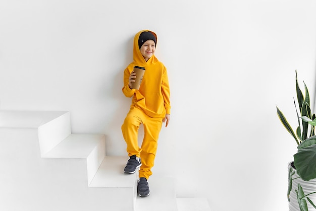 a teenager in a yellow tracksuit on a monochrome background in a black hat and hood with a drink