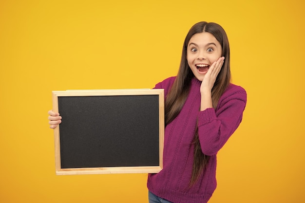 Teenager younf school girl holding school empty blackboard isolated on yellow background Portrait of a teen female student Excited face Amazed expression cheerful and glad