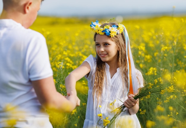 Teenagers brother and sister with Ukrainian wreath with on head in rapeseed field under blue sky