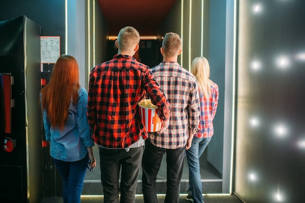 Teenagers with popcorn stands in cinema hall before the screening, back view. male and female youth in movie theater