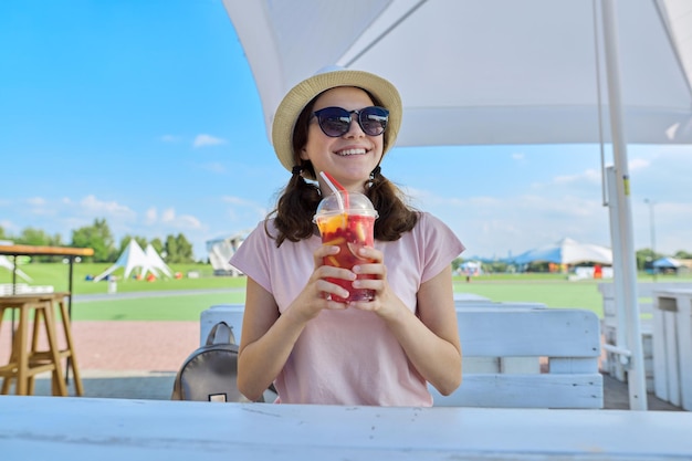 Teens, lifestyle, summer, vacation. Girl in outdoor cafe in hat sunglasses with berry drink.