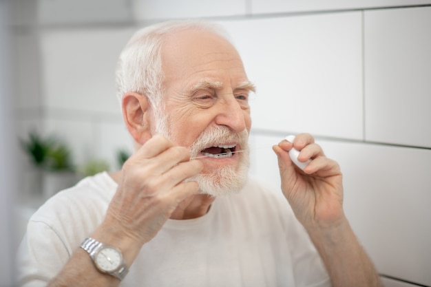 Pulizia dei denti. uomo dai capelli grigi in maglietta bianca che pulisce i denti con un filo interdentale