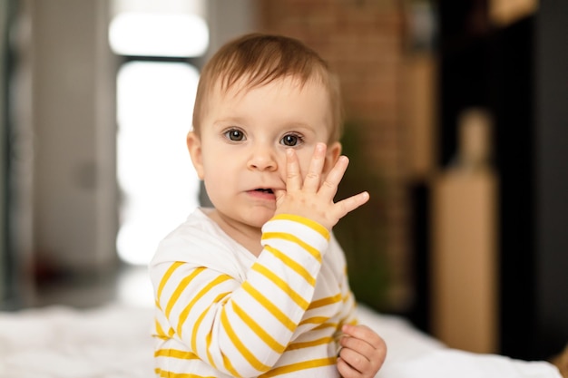 Teething concept Portrait of little baby girl sitting on white bedsheets and biting finger in mouth looking at camera