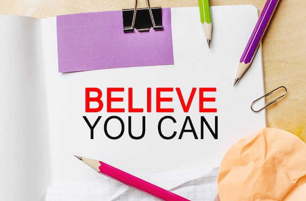 Tekst Believe you can op een witte notitie-achtergrond met potloden, stickers en paperclips
