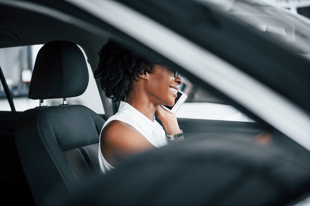 Telefoon gebruiken. Jonge Afro-Amerikaanse vrouw zit binnenkant van nieuwe moderne auto.