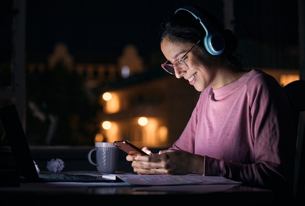 Telefoonnacht en vrouw netwerken met koptelefoon terwijl ze naar muziek, radio of podcast luistert Gelukkige glimlach en meisje browsen op sociale media mobiele app of internet met mobiel aan een bureau in haar huis