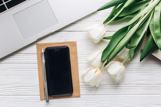 Telefoonscherm en laptop en tulpen op witte houten rustieke achtergrond stijlvolle flat lag met bloemen en werkende gadgets met ruimte voor tekst freelance thuiswerken