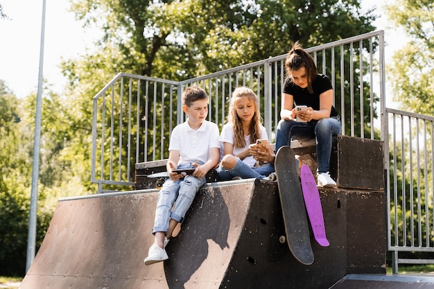 Telefoonverslaafde sportkinderen met skateboard en pennyboards zitten en kijken naar smartphones op sporthelling op speelplaats Kinderen verslaving aan telefoons