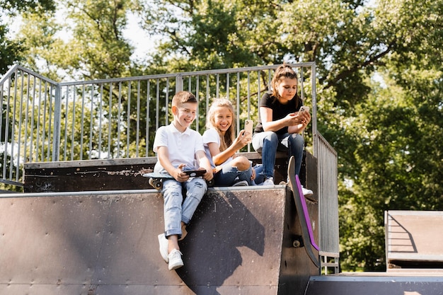Telefoonverslaafde sportkinderen met skateboard en pennyboards zitten en kijken naar smartphones op sporthelling op speelplaats Kinderen verslaving aan telefoons