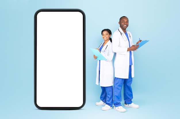 Telehealth-concept gelukkig mannelijke en vrouwelijke artsen in uniform staande in de buurt van enorme lege smartphone mockup