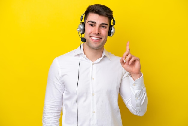 Telemarketeer blanke man aan het werk met een headset geïsoleerd op een gele achtergrond die een vinger toont en optilt in teken van de beste