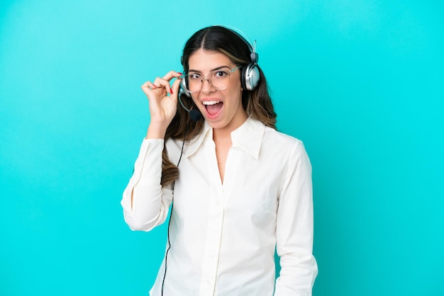 Telemarketeer Italiaanse vrouw die werkt met een headset geïsoleerd op een blauwe achtergrond met een bril en verrast