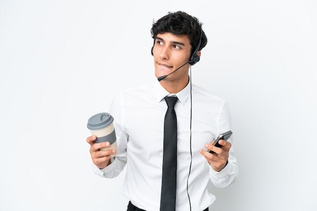 Telemarketeer man aan het werk met een headset geïsoleerd op een witte achtergrond met koffie om mee te nemen en een mobiel terwijl hij iets denkt