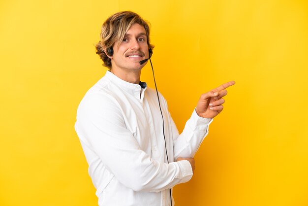 Telemarketeer man aan het werk met een headset geïsoleerd op gele muur wijzende vinger naar de zijkant