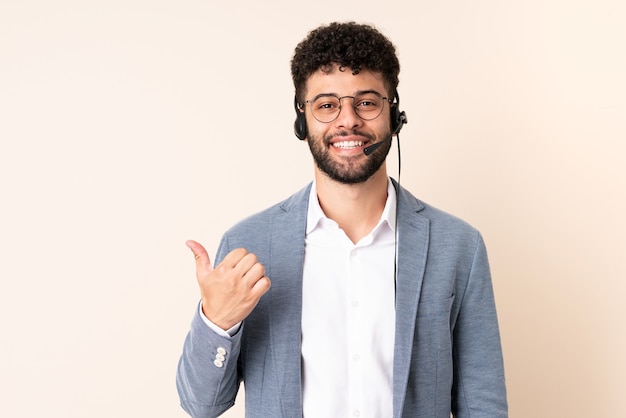 Telemarketeer Marokkaanse man aan het werk met een hoofdtelefoon geïsoleerd op beige muur wijzend naar de zijkant om een product te presenteren