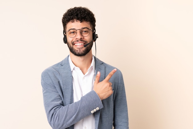 Telemarketeer Marokkaanse man aan het werk met een hoofdtelefoon geïsoleerd op een beige achtergrond wijst naar de zijkant om een product te presenteren