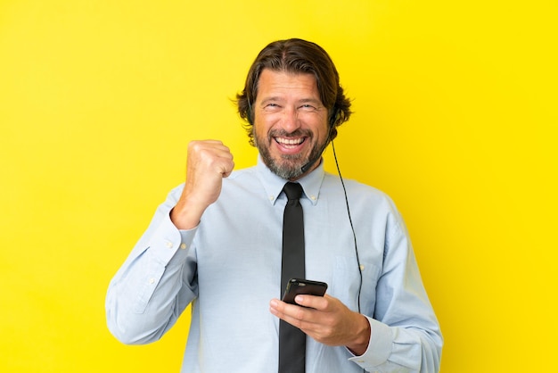 Telemarketeer Nederlandse man aan het werk met een headset geïsoleerd op gele achtergrond met telefoon in overwinningspositie