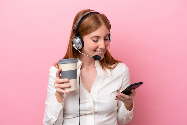 Telemarketeer roodharige vrouw die werkt met een headset geïsoleerd op roze achtergrond met koffie om mee te nemen en een mobiel