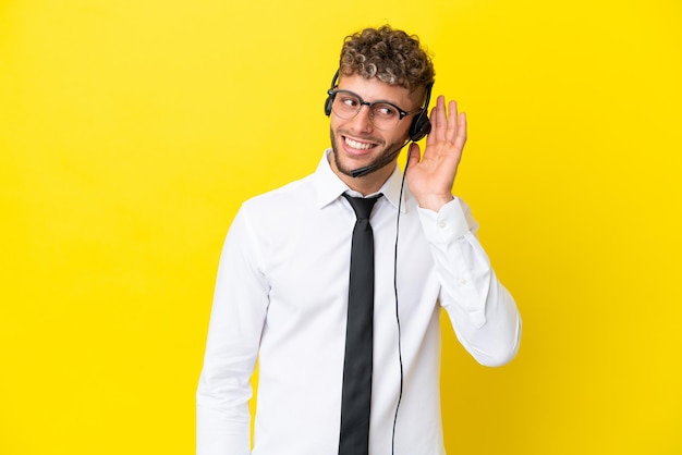 Telemarketer blonde man working with a headset isolated on yellow background listening to something by putting hand on the ear