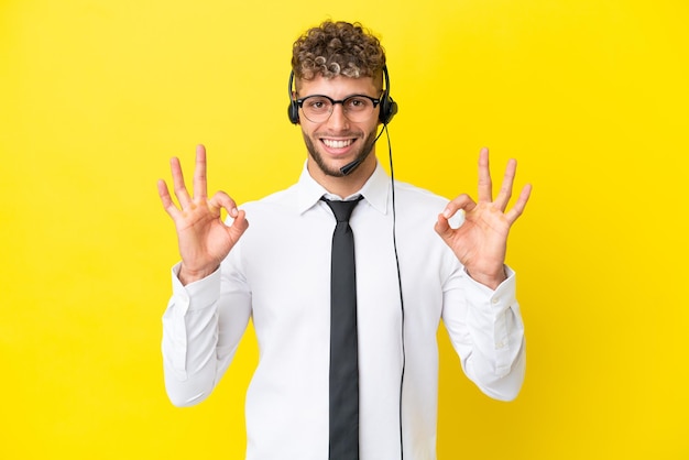 Telemarketer blonde man working with a headset isolated on yellow background showing an ok sign with fingers