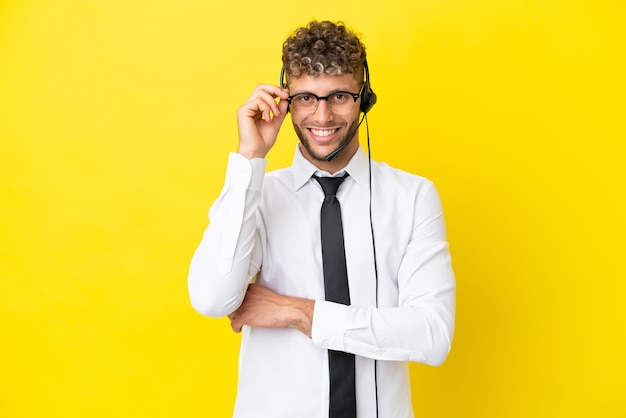 Telemarketer blonde man working with a headset isolated on yellow background with glasses and happy