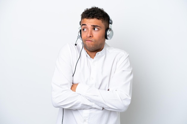 Telemarketer Brazilian man working with a headset isolated on white background making doubts gesture while lifting the shoulders