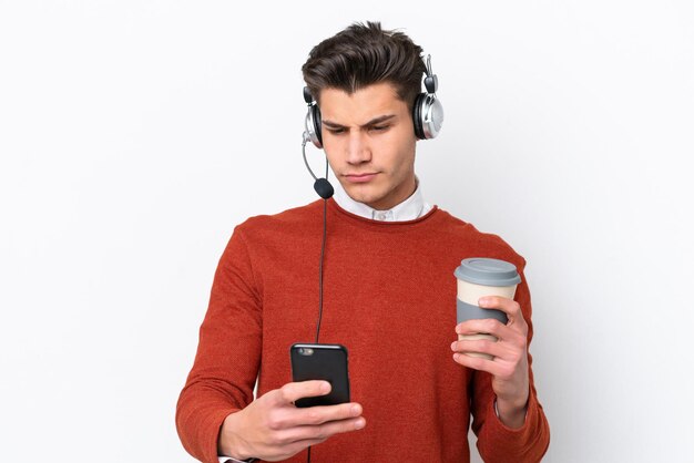 Telemarketer caucasian man working with a headset isolated on white background holding coffee to take away and a mobile