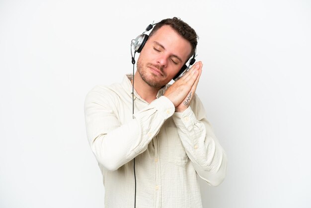 Telemarketer caucasian man working with a headset isolated on white background making sleep gesture in dorable expression