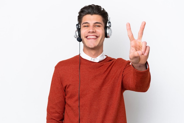 Telemarketer caucasian man working with a headset isolated on white background smiling and showing victory sign