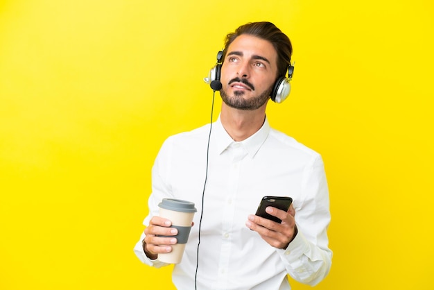 Telemarketer caucasian man working with a headset isolated on yellow background holding coffee to take away and a mobile while thinking something