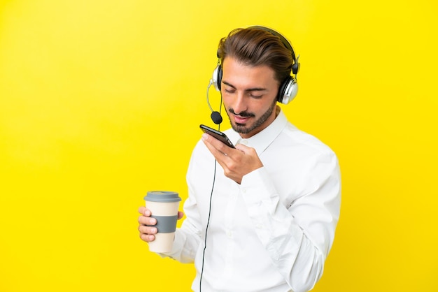 Telemarketer caucasian man working with a headset isolated on yellow background holding coffee to take away and a mobile