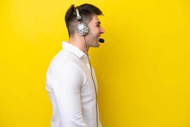 Telemarketer caucasian man working with a headset isolated on yellow background laughing in lateral position