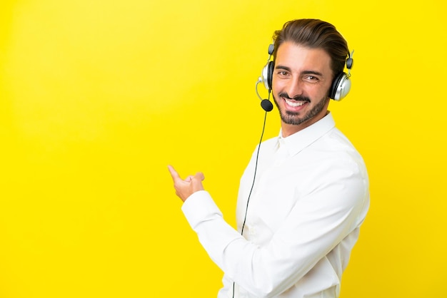 Telemarketer caucasian man working with a headset isolated on yellow background pointing back