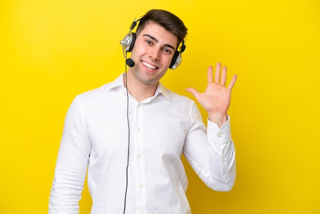 Telemarketer caucasian man working with a headset isolated on yellow background saluting with hand with happy expression