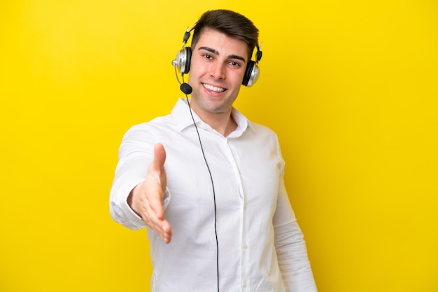 Telemarketer caucasian man working with a headset isolated on yellow background shaking hands for closing a good deal