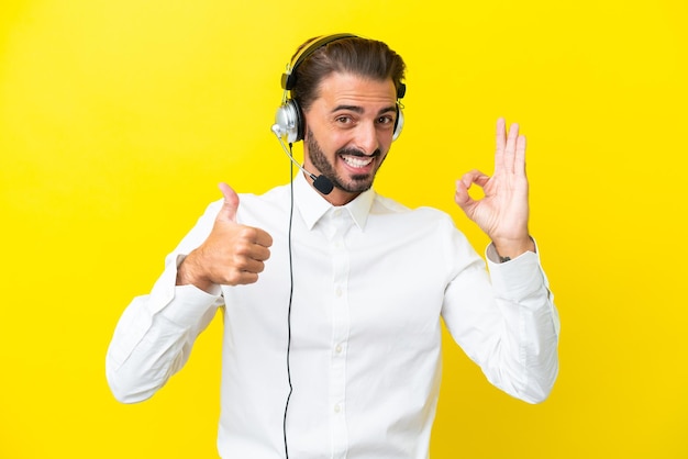 Telemarketer caucasian man working with a headset isolated on yellow background showing ok sign and thumb up gesture