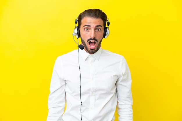 Telemarketer caucasian man working with a headset isolated on yellow background with surprise facial expression