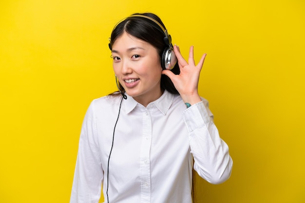 Telemarketer Chinese woman working with a headset isolated on yellow background listening to something by putting hand on the ear