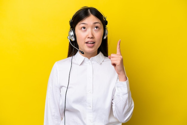 Telemarketer chinese woman working with a headset isolated on yellow background pointing up and surprised