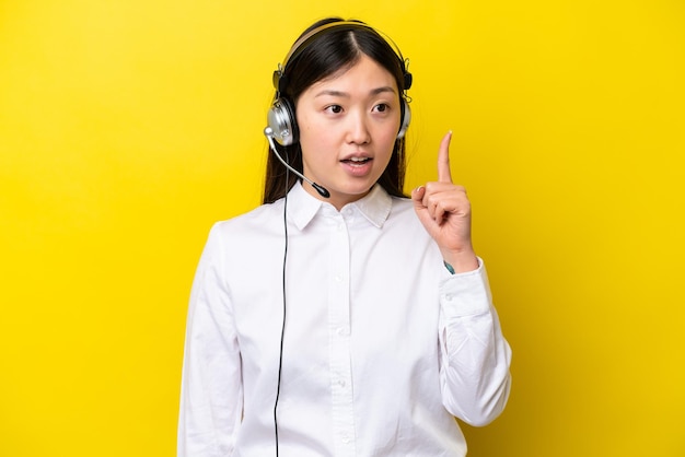 Telemarketer Chinese woman working with a headset isolated on yellow background thinking an idea pointing the finger up