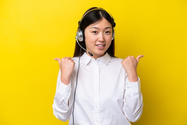 Телемаркетер китаянка, работающая с гарнитурой, изолированной на желтом фоне, с поднятым вверх пальцем и улыбкой