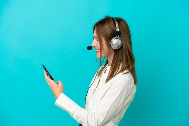 Телемаркетер англичанка, работающая с гарнитурой, изолированной на синем фоне, ведет с кем-то разговор по мобильному телефону