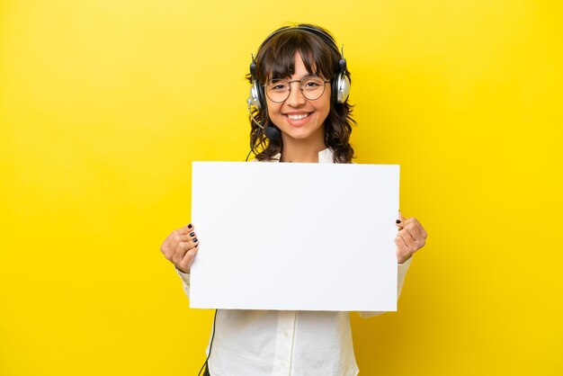 Telemarketer latin woman working with a headset isolated on yellow background holding an empty placard with happy expression