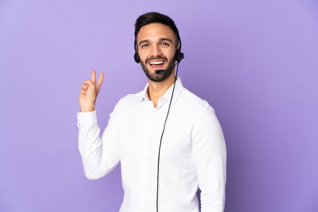 Telemarketer man working with a headset isolated on purple wall smiling and showing victory sign