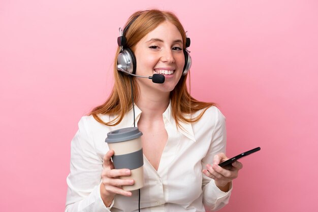 Telemarketer redhead woman working with a headset isolated on pink background holding coffee to take away and a mobile