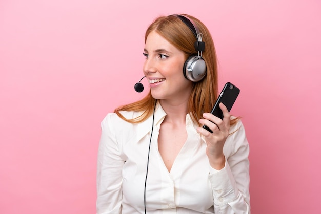 Telemarketer donna rossa che lavora con un auricolare isolato su sfondo rosa mantenendo una conversazione con il telefono cellulare con qualcuno